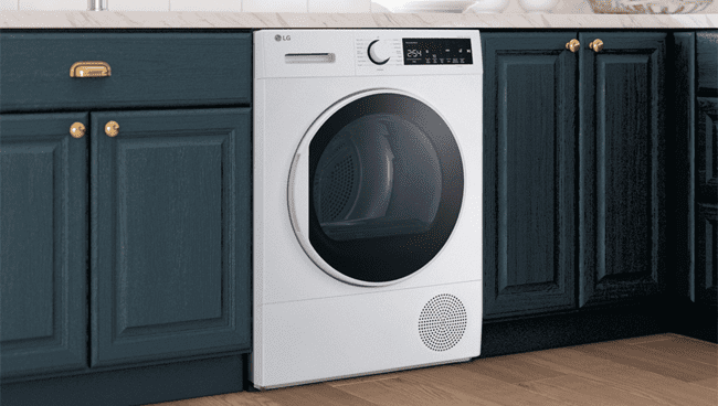 Image shows a white LG tumble dryer fitted into a modern kitchen with dark green/blue cabinets and white marble counter tops
