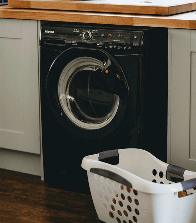 A image of a black freestanding washing machine in a traditional kitchen with a laundry basket of clothes in front of it.