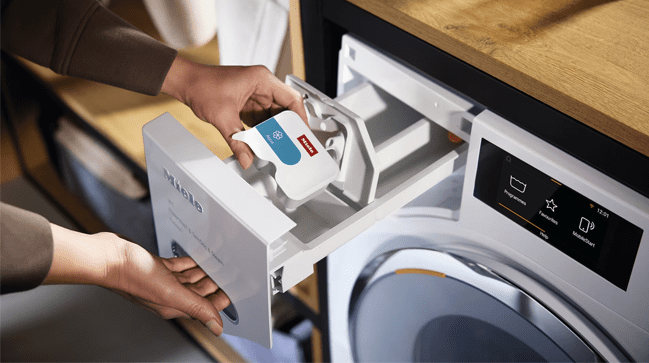 This image shows a hand reaching into the open top drawer of a Miele W1 washing machine holding onto a capdosing capsual