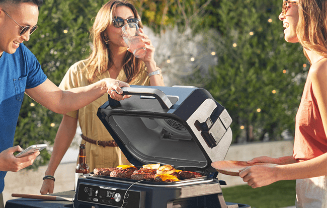 A group of friends laugh and enjoy drinks around a Ninja BBQ grill- now available at appliance city along with other Ninja outdoor cooking appliances.