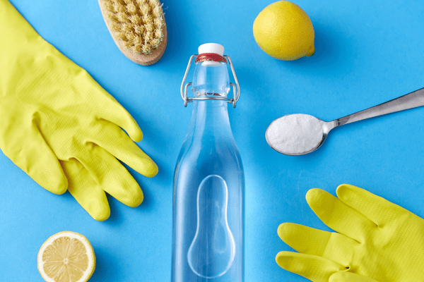 Image of cleaning products to clean a kitchen sink. These include yellow washing up gloves, a teaspoon of bicarbonate of soda, a bottle of white vinegar, some lemons and a bristle brush. All on a blue background.