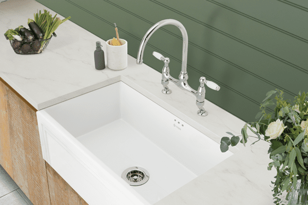 Lifestyle image of a ceramic Caple Belfast sink. Set into a white kitchen worktop with light wooden cabinets and sage green panelling behind.