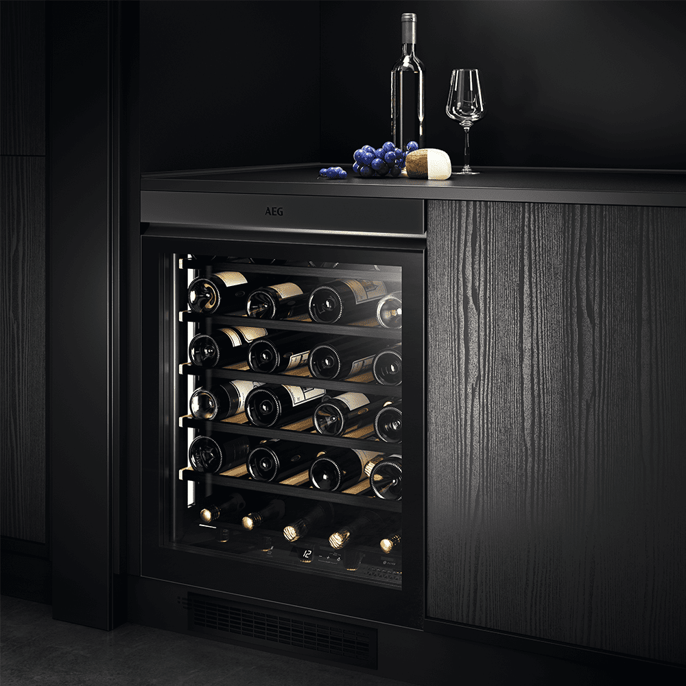 Image of an AEG undercounter wine cooler, positioned under a dark grey wooden kitchen. Above the wine cooler is a bottle of wine with a glass and some grapes