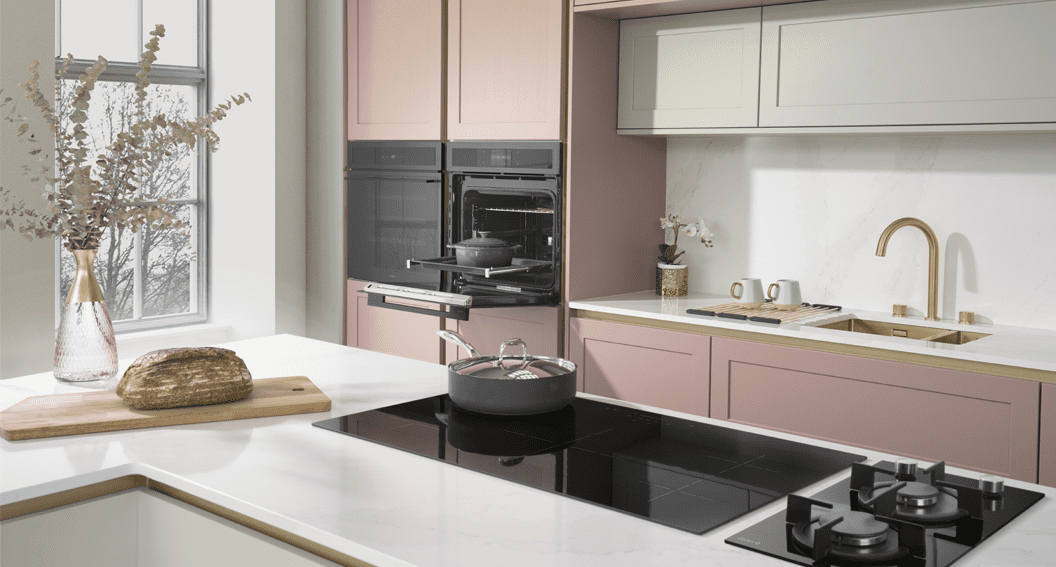 Image of a pink Caple kitchen featuring gold details. In the kitchen you can see a gold Caple tap and sink, two built in ovens, an induction hob and a small gas domino hob.
