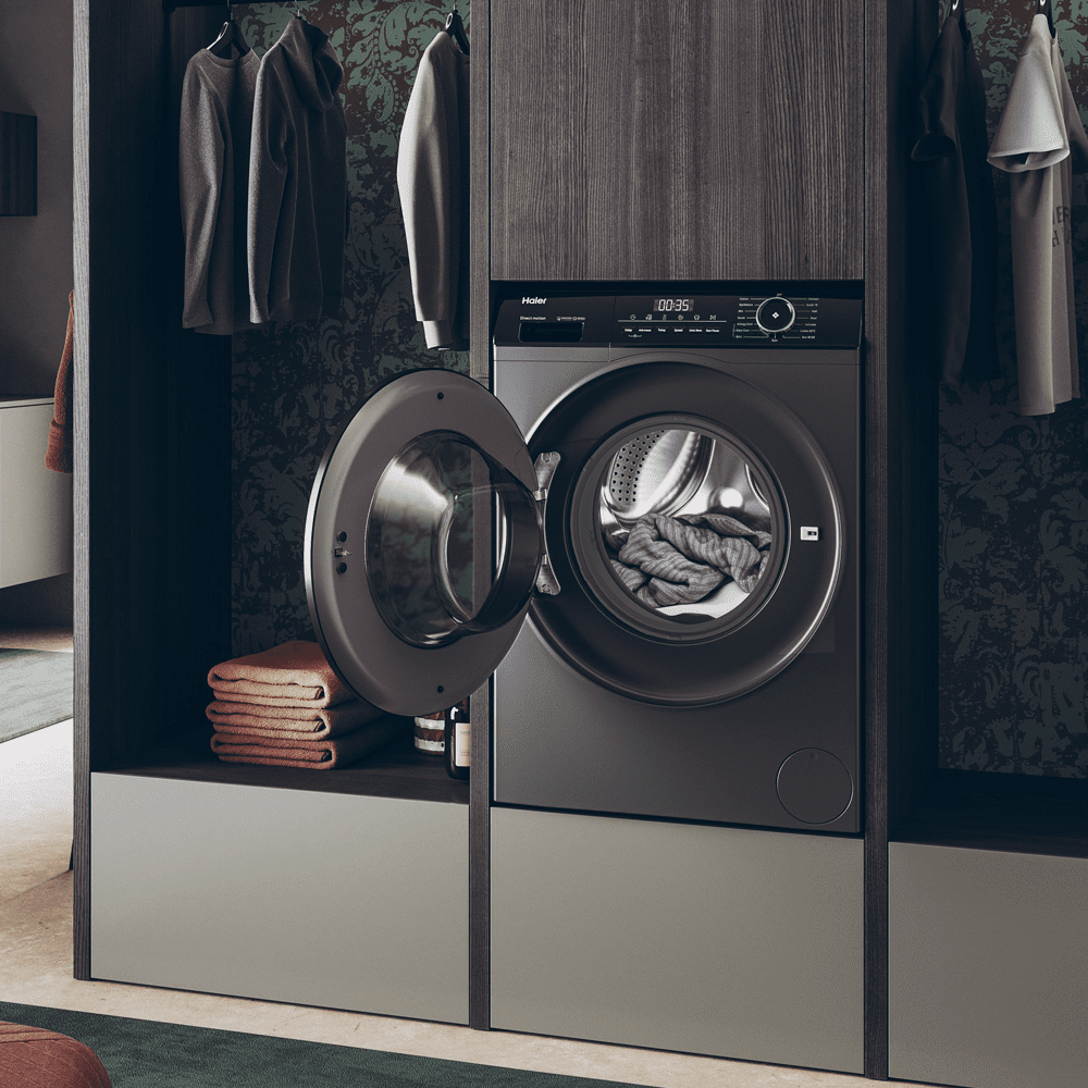 A Haier washing machine finished in graphite positioned in a dark wooden utility room. The washing machine door is open, with the interior of the appliance visible.