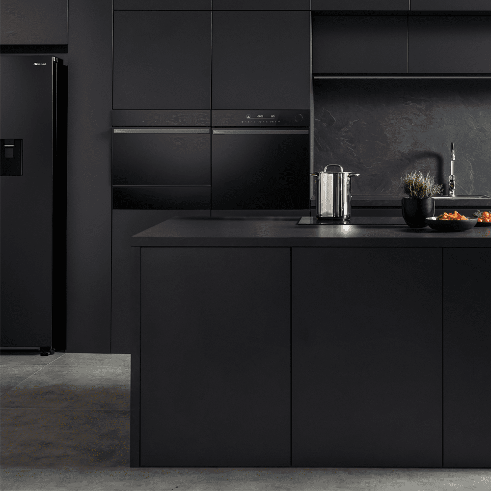 A dark and moody lifestyle image of a black kitchen featuring a built in oven, microwave and induction hob.