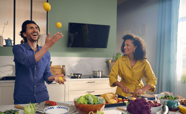 A couple standing in their kitchen in front of their NEFF cooker hood, preparing dinner. The man is juggling lemons whilst the woman is preparing vegetables.