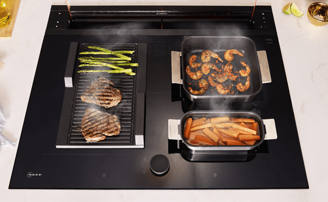 Close up of a NEFF induction hob featuring sizzling steaks, vegetables and prawns.