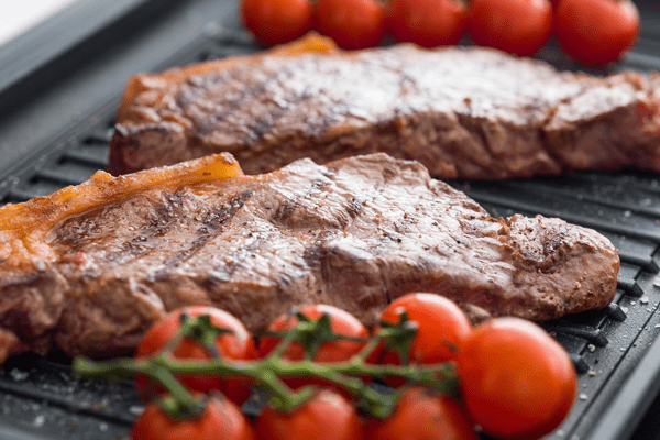 A close up of an AGA Masterchef griddle. The griddle features cherry tomatoes on the vine cooking alongside a sizzling steak.
