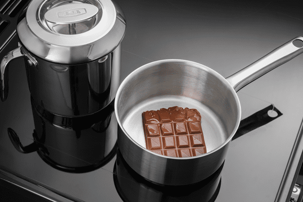 A close up image of an AGA Masterchef induction range cooker. The image features two pans on the hob top, including one saucepan which is melting chocolate.