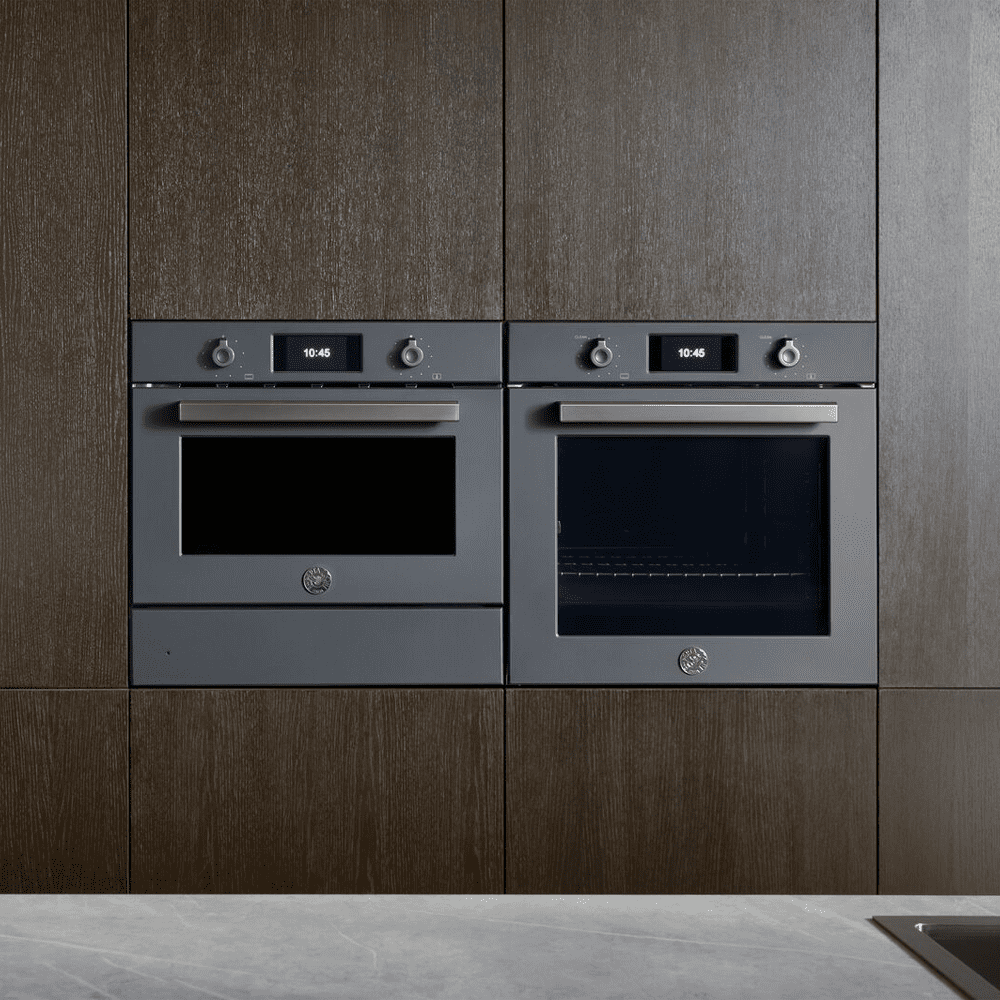 Image of a built in oven and combination microwave and warming drawer sat alongside each other. Built into a dark wooden tall housing unit, the Bertazzoni appliances are finished in grey.