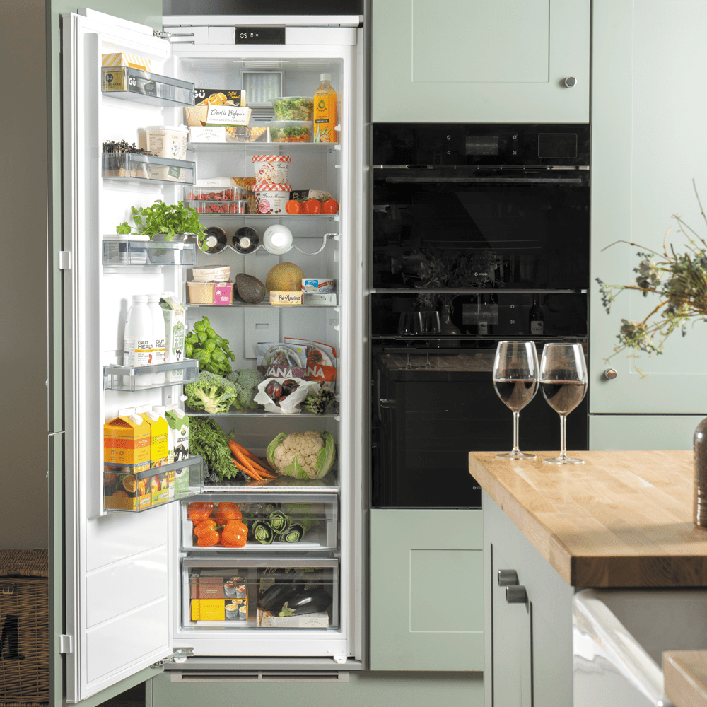 Image of a Caple fridge in a modern, mint green kitchen. The fridge door is open and you can see brighlty coloured fresh fruit and vegetables on the shelves. Next to the fridge are two Caple built in ovens.