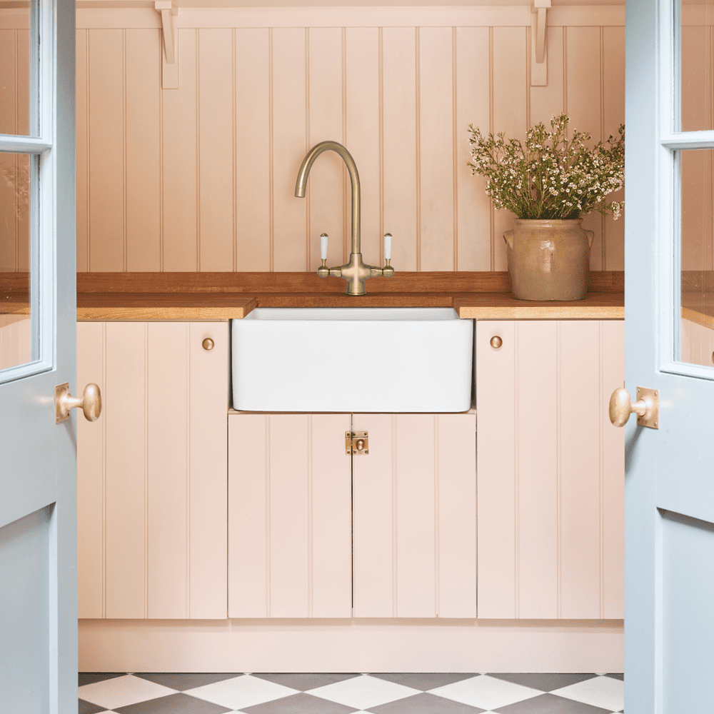 Lifestyle image of a pastel peach coloured utility room featuring a Caple Belfast sink in white, with a gold tap.