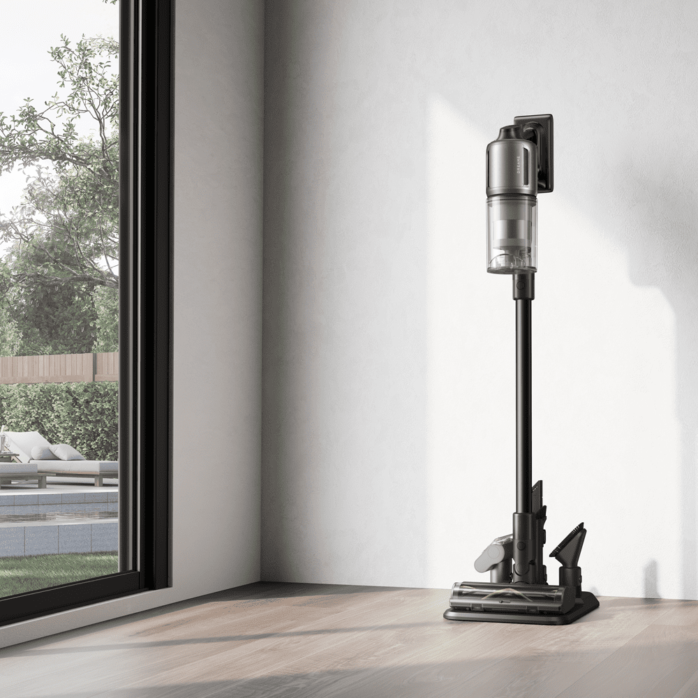 Lifestyle image of a Dreame cordless stick vacuum cleaner finished in black, standing upright in the corner of a light grey room.