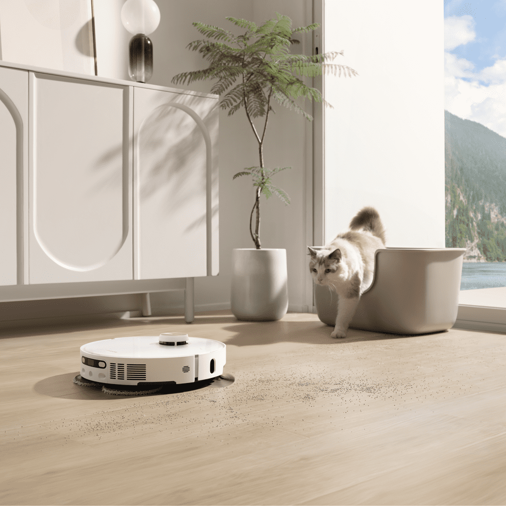 image of a Dreame robot vacuum cleaner finished in white, on a hard wooden floor cleaning up debris. Behind the vacuum cleaner, there is a cat moving towards the appliance.