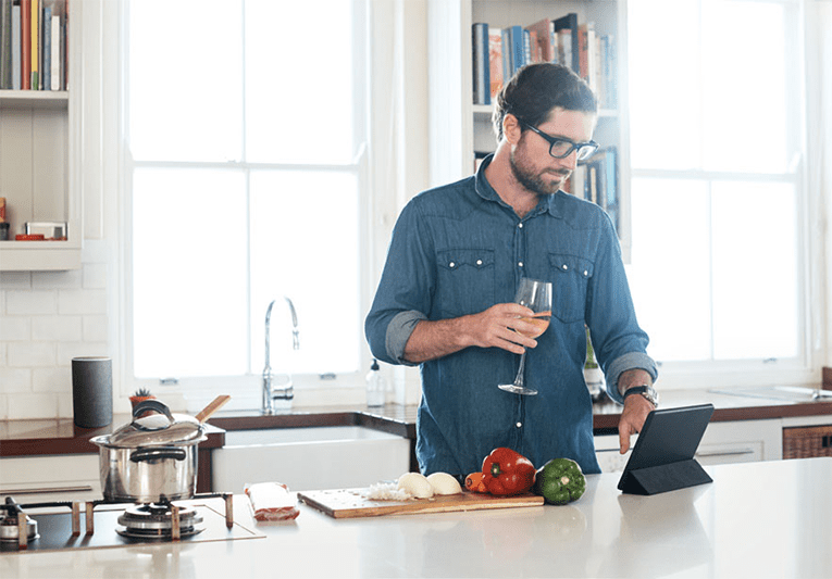 Image of a man reading a recipe off a smart device, drinking a glass of wine whilst he is cooking