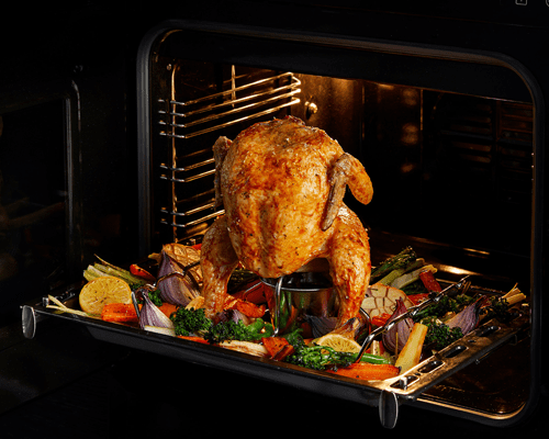 Close up of a roast chicken in a Stoves range cooker, on a rotisserie with brightly coloured vegetables beneath it in the pan.