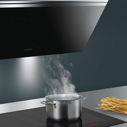 Close up of a Siemens angled chimney hood in black, positioned over an induction hob. A pan is boiling on the hob and you can see the steam rising into the extractor.