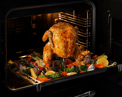 Close up of a chicken inside a Stoves range cooker, cooking on a rotisserie with vegetables on the tray beneath it.