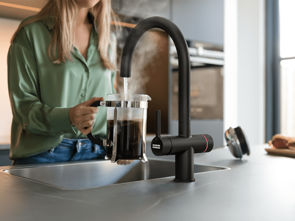 Close up of a woman pouring boiling water into a cafetiere in a modern grey kitchen. The Franke tap is an electronic Water Hub finished in matte black.