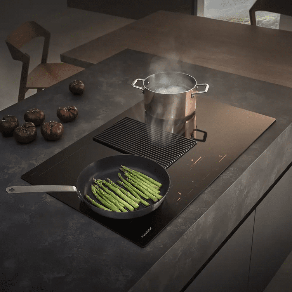 Close up of a Samsung air venting induction hob in black. On the hob top there is a pan of boiling water and a frying pan filled with asparagus