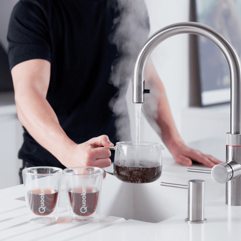 Close up of a Quooker boiling water tap in use. Two cups of coffee are sat beside the sink and a man is using the tap to pour boiling water into a carafe.