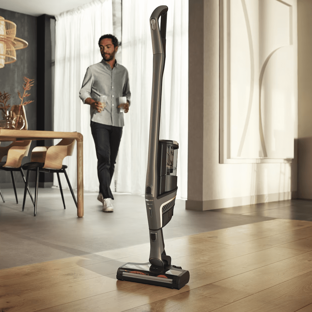 Image of a Miele cordless vacuum cleaner standing on a hard wooden floor in an open plan kitchen dining room.