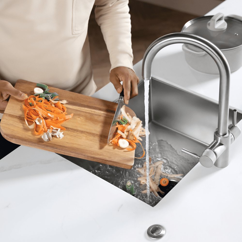 Close up of a waste disposal unit in use. Someone is scraping vegetable peelings into the sink along with water from the tap. This then filters through the plug hole and is ground down into fine particles that can be washed down the waste pipe.